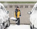 Snow falls heavily. A man in a yellow and black coat stands between two parked cars holding a big shovel full of snow.
