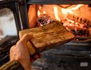 A hand holding a log reaches toward a fireplace.