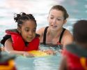 A child in a life jacket swims on a paddle board with an adult and other kids nearby.