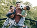 A dad laughs while playing outside with two laughing kids.