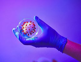 A hand wearing a medical glove holding an illustration of the COVID-19 virus.