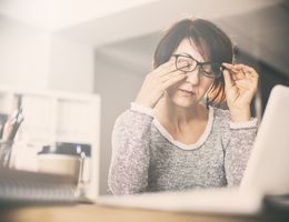 A woman rubs her eyes while working at a laptop