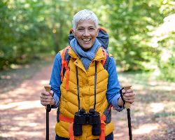 An older woman with hiking poles stands on a trail, smiling.