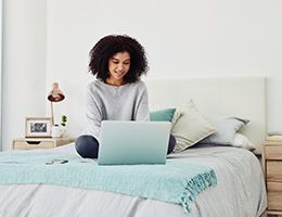 A woman sits on her bed with a laptop.