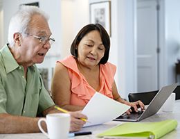 An older couple sit at a laptop and look at paperwork.