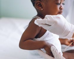 A toddler sits on an exam table as a gloved hand cleans her shoulder.