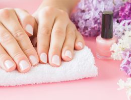 A pair of manicured hands resting on a towel next to nail polish and lilacs.
