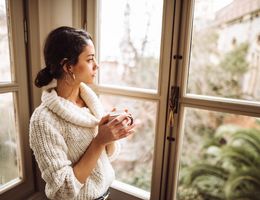 A young woman looks pensively out a window, holding a mug in her hands.