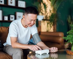 A man sits on a couch and uses a blood pressure monitor.