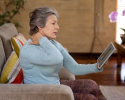 An older woman reaches to her shoulder with one hand while the other holds a tablet.