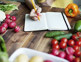 A hand makes notes in a notebook on a produce-covered table.