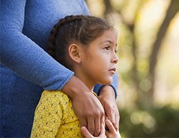 A mom is standing behind a young girl with her arms resting on the girl's shoulders.