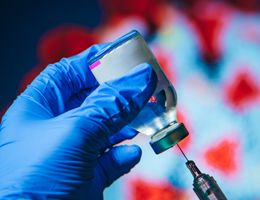 A gloved hand draws vaccine into a syringe from a vial in front of an image of the coronavirus.