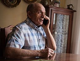 An older man sits at a table and talks on a phone.