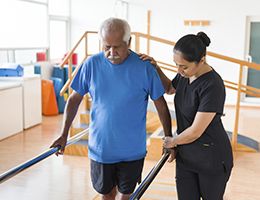 A physical therapist helps a man walk between parallel bars.