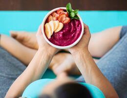 A woman, seen from above, sits cross-legged while holding a smoothie bowl topped with strawberry and banana slices.