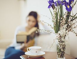 A woman is sitting alone reading a book. A tea cup and flowers are nearby.