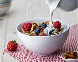 Milk pours from a pitcher onto a bowl of cereal topped with fruit.