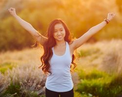 A smiling young woman in a tank top stands in a sunlit field, arms raised in a wide pose