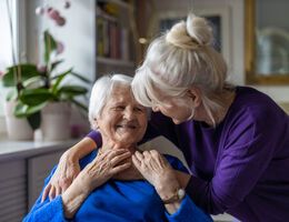 A woman bends to hug a seated, smiling older woman