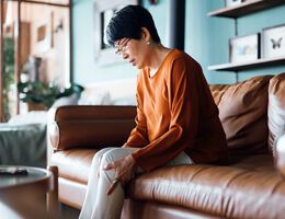 A woman seated on a couch massages her knee with both hands.