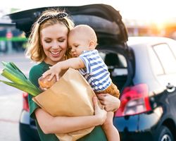 A woman holds a baby and a bag of groceries. Her car is behind her with the trunk open.