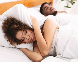Snoring man lying next to grimacing woman with pillow folded over her ears.