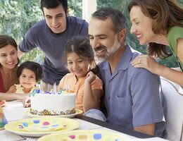 A family gathers around as a man blows out birthday candles on a cake