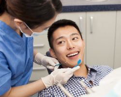 A dental hygienist holds a pick and mirror near a smiling young man in a dental chair