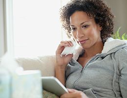 A woman holding a tissue sits on a couch and looks at a laptop.
