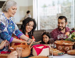 A multigenerational family sit down for a meal as an older woman adds another dish to the table.