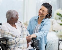 A young woman holds the hand of an older woman wrapped in a blanket and seated in a wheelchair.