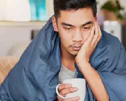 A man wrapped in a blanket holds a mug and rests his head on his hand.