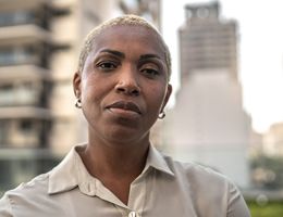 A woman with a serious expression stands in front of urban buildings.