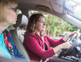 A young woman driving a car as an older passenger watches