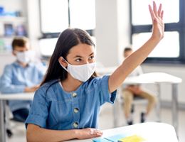 A girl wearing a face mask raises her hand in a classroom.