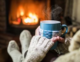 View of hands holding a mug as feet are stretched toward a fireplace