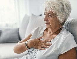 A woman sits on a couch with a hand pressed to her chest