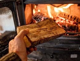 A hand holding a log reaches toward a fireplace.