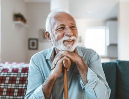 An older man sitting on a couch with his hands on his cane and his head leaning on his hands.