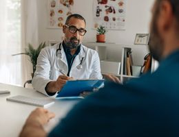 A doctor holds a clipboard while talking across his desk to a patient