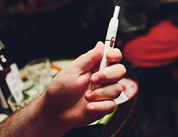 A hand holds a heat-not-burn device in front of a dining table.