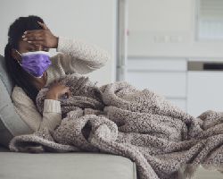A masked woman lies on a couch, one hand to her forehead.