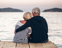 An older couple sits on a dock, his arm around her shoulders