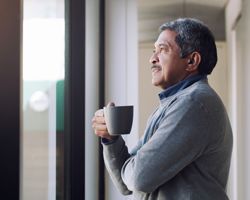 A man holds a mug and looks out a window.