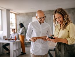 A man and a woman stand in an open office, smiling as they look at her phone