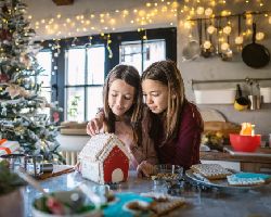Two young girls decorate a gingerbread house.