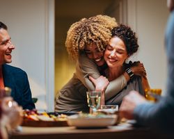A woman hugs another woman who is seated at a table.