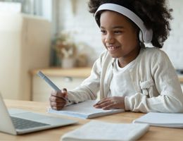 A smiling girl at a table with headphones and a laptop.