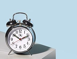 A silver alarm clock sits on a gray table in front of a blue wall